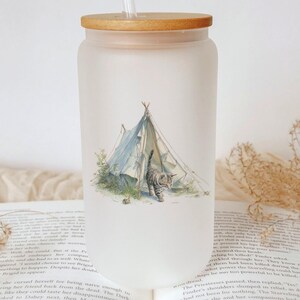 May include: Frosted glass tumbler with a wooden lid and straw. The tumbler features a watercolor illustration of a tent and a tabby cat. The design is in soft blues and greens, evoking a camping theme.