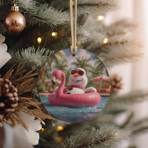 May include: Round Christmas ornament featuring Olaf from Frozen, wearing sunglasses and a scarf, floating in a pink flamingo pool float. The background shows a tropical pool scene. The ornament hangs from a gold string.