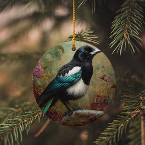 May include: Round Christmas ornament featuring a detailed painting of a magpie bird with black, white, and teal plumage. The bird is perched on a branch against a backdrop of floral elements. A gold string is attached for hanging.