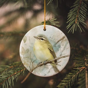 May include: A round ceramic ornament featuring a watercolor painting of a yellow and gray bird perched on a branch. The bird is set against a soft, muted background with subtle leaf details. A gold string is attached for hanging.