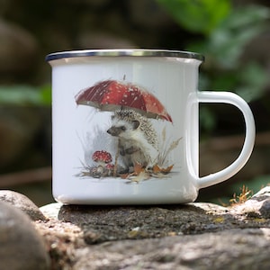 May include: White enamel mug with a whimsical illustration of a hedgehog sheltering under a red and white mushroom umbrella. The mug has a silver rim and a white handle, perfect for coffee or tea.