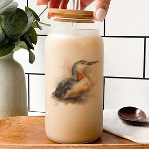May include: A glass tumbler with a bamboo lid and straw, featuring a vintage illustration of a bird. The tumbler is filled with a light-colored beverage and sits on a wooden tray. A wooden spoon and a white cloth are also visible.
