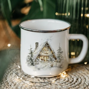 May include: White enamel mug with a winter cabin scene. The mug features a black rim and a handle. The cabin is surrounded by snow-covered trees and is lit up with warm lights, creating a cozy, festive feel. The mug sits on a woven surface.