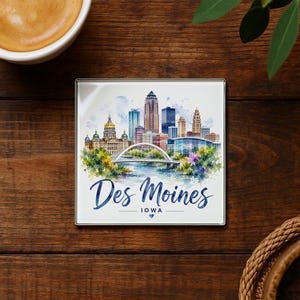 May include: Square coaster featuring a watercolor illustration of the Des Moines, Iowa skyline. The design includes the city's buildings, a bridge, and the text "Des Moines Iowa" in a script font. The coaster has a silver border.