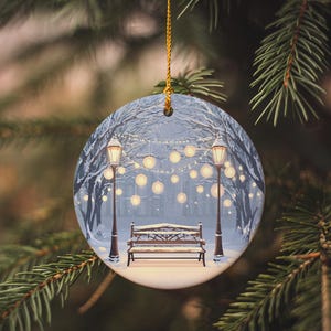 May include: Round Christmas ornament featuring a winter scene with a snow-covered bench between two lampposts. The ornament has a gold string for hanging and is set against a backdrop of evergreen branches.