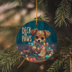 May include: Round Christmas ornament with a teal background, featuring a cute puppy tangled in colorful Christmas lights. The text "Deck the Paws" is printed on the ornament. The ornament is hanging from a gold string against a backdrop of pine needles.
