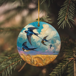 May include: Round ornament featuring a painting of swallows in flight against a cloudy sky and wheat field. The birds are blue and black with white accents. The ornament is hanging from a gold string.