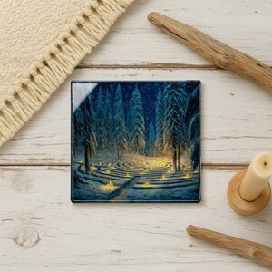 May include: Square glass coaster featuring a winter forest scene with a candlelit labyrinth. The artwork depicts tall, snow-covered trees under a dark blue sky. The coaster is placed on a white wooden surface with decorative elements.