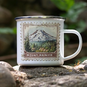 May include: White enamel mug featuring a vintage-style Mount Rainier design. The mug has a silver rim and a curved handle. The design depicts a mountain landscape with trees and the words "Mount Rainier" in a brown frame. The mug is sitting on a rock.
