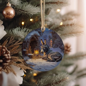May include: Round Christmas ornament featuring a winter scene with a donkey resting near a lantern. The ornament has a gold string for hanging and is displayed on a Christmas tree with other ornaments and lights.