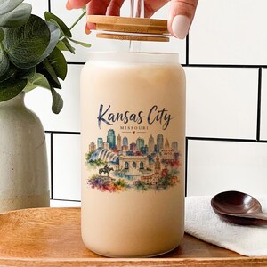 A hand is opening a large glass jar with a label featuring a cityscape, likely representing Kansas City, Missouri. The jar is placed on a wooden tray, and there are some decorative elements in the background, such as a potted plant and a bottle.
