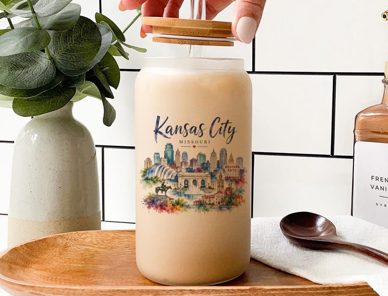 A hand is opening a large glass jar with a label featuring a cityscape, likely representing Kansas City, Missouri. The jar is placed on a wooden tray, and there are some decorative elements in the background, such as a potted plant and a bottle.