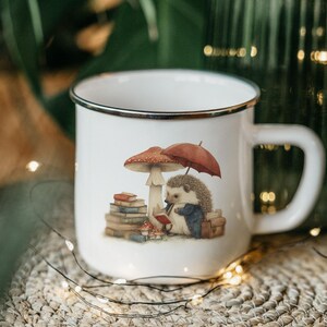 May include: White enamel mug with a black rim featuring a whimsical illustration of a hedgehog reading under a mushroom and umbrella. The mug has a large handle and is set on a woven surface.