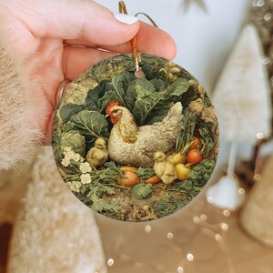 May include: Round ceramic ornament featuring a hen with chicks amidst a garden of green leafy vegetables and orange carrots. The ornament has a gold string for hanging. The artwork has a vintage, rustic style.