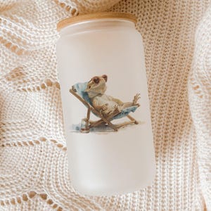 May include: Frosted glass tumbler with a bamboo lid. The tumbler features a whimsical illustration of a frog wearing sunglasses, relaxing in a beach chair. The frog is enjoying a drink. The tumbler is resting on a cream-colored knitted blanket.