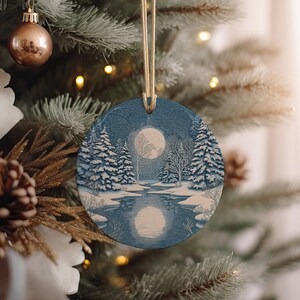 May include: Round Christmas ornament with a winter landscape scene. The ornament features a full moon, snow-covered trees, and a reflective body of water. The ornament is blue and white, with a gold string for hanging. The ornament is hanging on a Christmas tree.