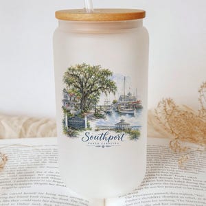A white ceramic mug with a printed design of a coastal scene, featuring a tree, boats, and a lighthouse, sits on top of an open book.