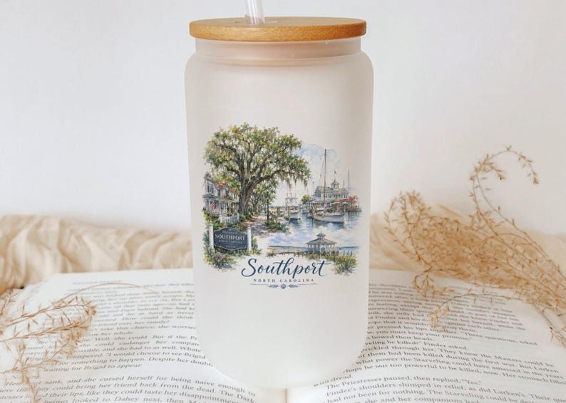 A white ceramic mug with a printed design of a coastal scene, featuring a tree, boats, and a lighthouse, sits on top of an open book.