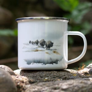 May include: White enamel mug with a silver rim and a white handle. The mug features a watercolor painting of a herd of bison walking across a snowy landscape. The mug is sitting on a stone surface.
