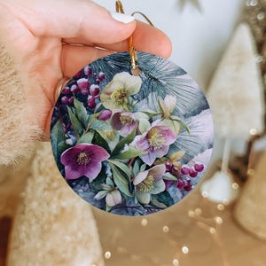 May include: Round Christmas ornament featuring a watercolor floral design with purple, pink, and yellow flowers, green leaves, and red berries. The ornament has a gold string for hanging and is a festive holiday decoration.