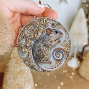 May include: A round Christmas ornament featuring a detailed illustration of a mouse. The mouse is depicted in shades of gray and gold, with intricate scrollwork and a curled tail. The ornament has a gold hanging loop.