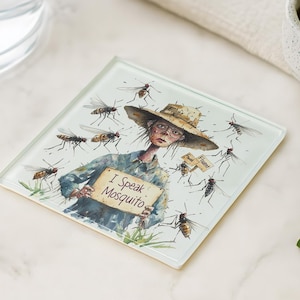 May include: Square glass coaster featuring a whimsical illustration of a person surrounded by mosquitoes. The person holds a sign that reads "I Speak Mosquito." The artwork is detailed with a straw hat and glasses. The coaster has a light green border.