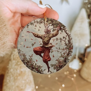 May include: Round ceramic ornament featuring a dancing figure with antlers, clad in a brown tunic and red pants, surrounded by foliage and falling leaves. The ornament has a gold string for hanging, perfect for holiday decor.