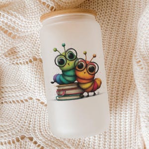 May include: Frosted glass tumbler with a bamboo lid. The tumbler features a colorful illustration of two cartoon caterpillars wearing glasses, sitting on a stack of books. The design has a whimsical, playful aesthetic.
