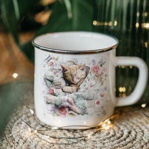 May include: White enamel mug with a silver rim, featuring a sleeping orange tabby kitten nestled in a floral quilt. The mug has a large handle and is decorated with pastel flowers and sewing notions.