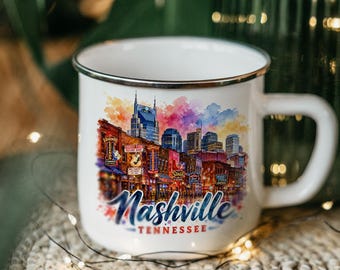 Könnte beinhalten: Ein weißer Emaillebecher mit einer farbenfrohen Aquarell-Illustration der Skyline und Straßenszene von Nashville, Tennessee. Das Design zeigt verschiedene Leuchtreklamen und den Schriftzug Nashville Tennessee in blauer Schreibschrift.