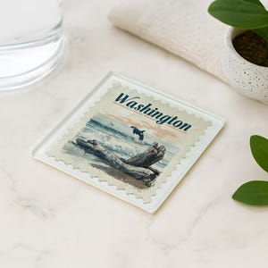May include: Square glass coaster with a vintage-style illustration of a Washington state coastal scene. The image features a breaching orca, driftwood, and ocean waves. The word "Washington" is printed across the top in a serif font.