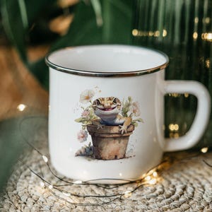 May include: White enamel mug with a black rim and handle. The mug features a watercolor illustration of a gecko in a flower pot with pink and white flowers. The mug is sitting on a woven surface.