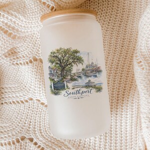 A frosted glass jar with a printed image of a coastal scene, including a tree, boats, and buildings, is placed on a white knitted blanket.