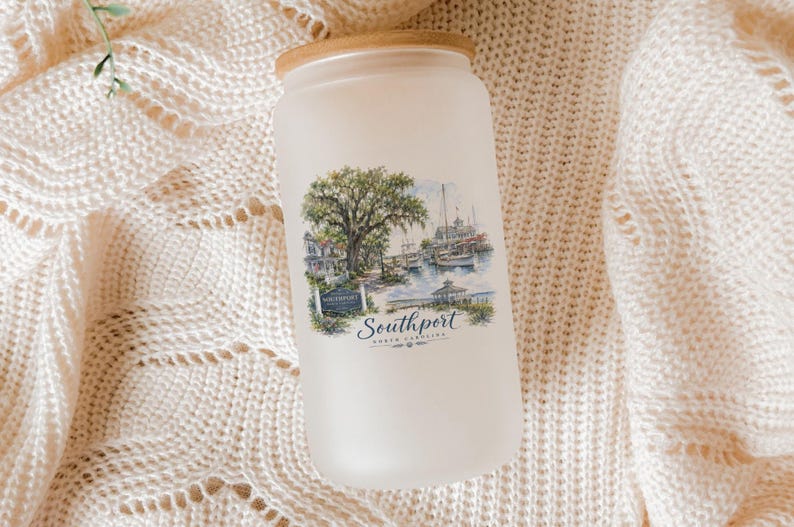 A frosted glass jar with a printed image of a coastal scene, including a tree, boats, and buildings, is placed on a white knitted blanket.