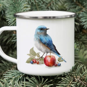 May include: White enamel mug with a detailed watercolor illustration of a bluebird perched on a branch with a red apple and berries. The mug has a silver rim and a white handle.