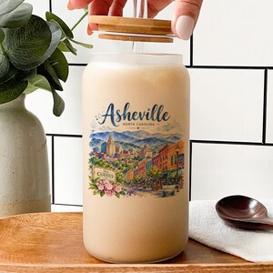 A hand is opening a large glass jar with a label that features an illustration of a cityscape, likely representing the city of Asheville, North Carolina. The jar is placed on a wooden tray, and there are some decorative elements in the background, such as a potted plant and a bottle.