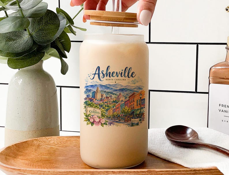 A hand is opening a large glass jar with a label that features an illustration of a cityscape, likely representing the city of Asheville, North Carolina. The jar is placed on a wooden tray, and there are some decorative elements in the background, such as a potted plant and a bottle.