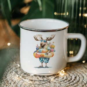 May include: White enamel mug with a black rim featuring a cartoon moose wearing sunglasses, a shirt, and swim trunks, carrying beach gear and a colorful inflatable ring. The mug has a large handle.