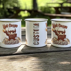 May include: Three white enamel mugs with a silver rim. The mugs feature a cartoon character holding a weapon and the text "Hot Cocoa Commando". One mug has the text "Just Us" on it.