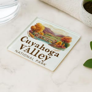 May include: Square glass coaster featuring a scenic illustration of Cuyahoga Valley National Park. The image includes a bridge, trees with fall foliage, and mountains. The text "Cuyahoga Valley National Park" is printed in brown.