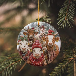 May include: A round Christmas ornament featuring a group of cows wearing festive attire. The cows are adorned with scarves, hats, and wreaths, set against a snowy backdrop. The ornament hangs from a gold string.