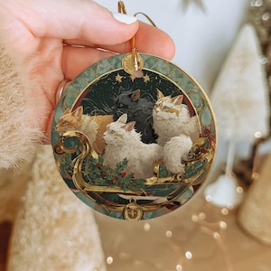 A hand holding a decorative ornament featuring a group of cats on a sleigh, with a starry night sky background.