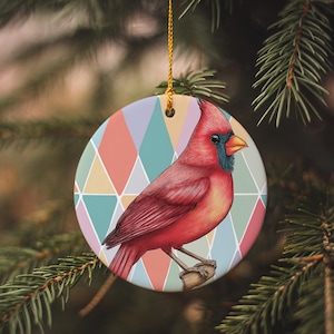 May include: A round ceramic ornament featuring a vibrant red cardinal perched on a branch. The bird is set against a backdrop of pastel-colored diamond shapes. A gold string is attached for hanging, perfect for holiday decor.
