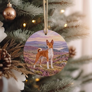 May include: A round ceramic ornament featuring a detailed illustration of a brown and white Basenji dog standing in a field of purple heather. The background shows rolling hills and a distant lake under a soft sunset sky. A gold cord is attached.