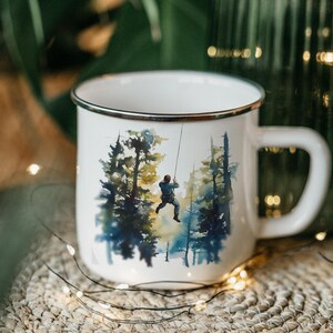 May include: White enamel mug with a black rim and a handle. The mug features a watercolor illustration of a person rappelling down a rope in a forest. The trees are painted in shades of blue, green, and yellow.