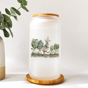 May include: A frosted glass tumbler with a bamboo lid and straw. The tumbler features a watercolor illustration of a duck in a bonnet walking through a garden of cabbages. The tumbler sits on a round bamboo coaster.