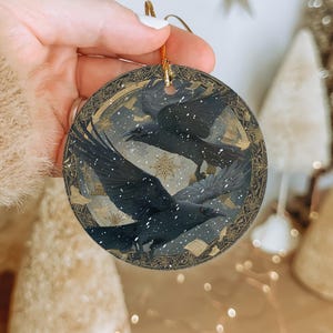 A hand is holding a circular ornament featuring a black bird with outstretched wings against a snowy background, with a star visible in the top right corner.