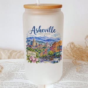 A white glass jar with a printed design of a cityscape and mountains, featuring the text "Asheville, North Carolina" and "Curiously", placed on top of an open book.