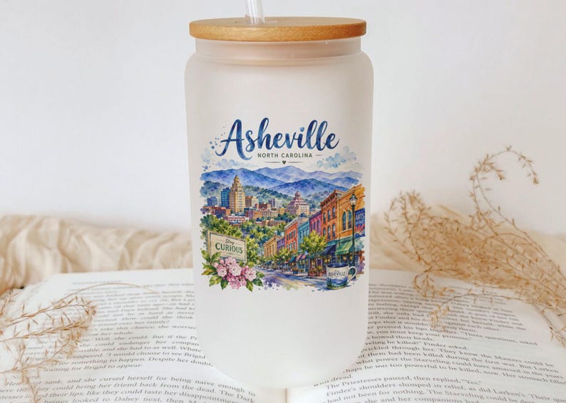 A white glass jar with a printed design of a cityscape and mountains, featuring the text "Asheville, North Carolina" and "Curiously", placed on top of an open book.