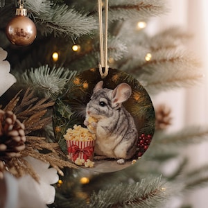 May include: Round Christmas ornament featuring a chinchilla eating popcorn. The chinchilla is gray and white, with large ears and a fluffy tail. A red and white striped popcorn container is next to the chinchilla. The ornament hangs from a gold string.